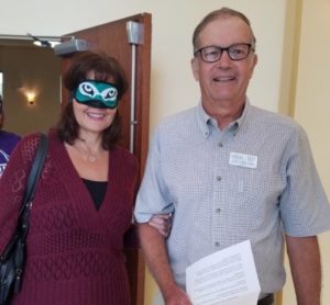 A person wearing a blindfold holds the arm of another. The person without a blindfold hold a printed page with an assignment on it. Both people are looking at the camera smiling.