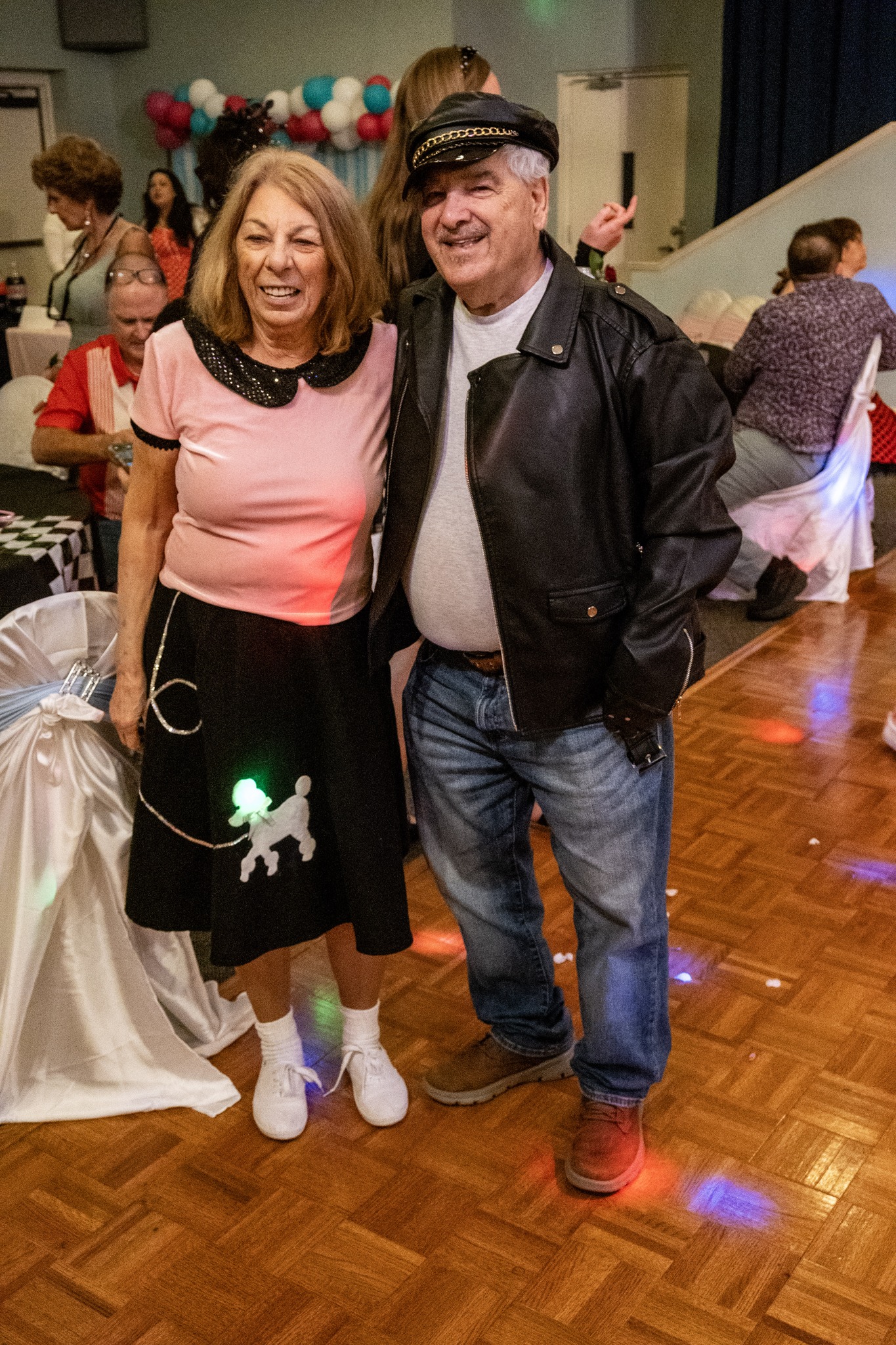 Two guests smile and pose together for a photo at the Senior Prom. One is wearing a pink shirt and a black poodle skirt, the other is in a leather jacket and hat.
