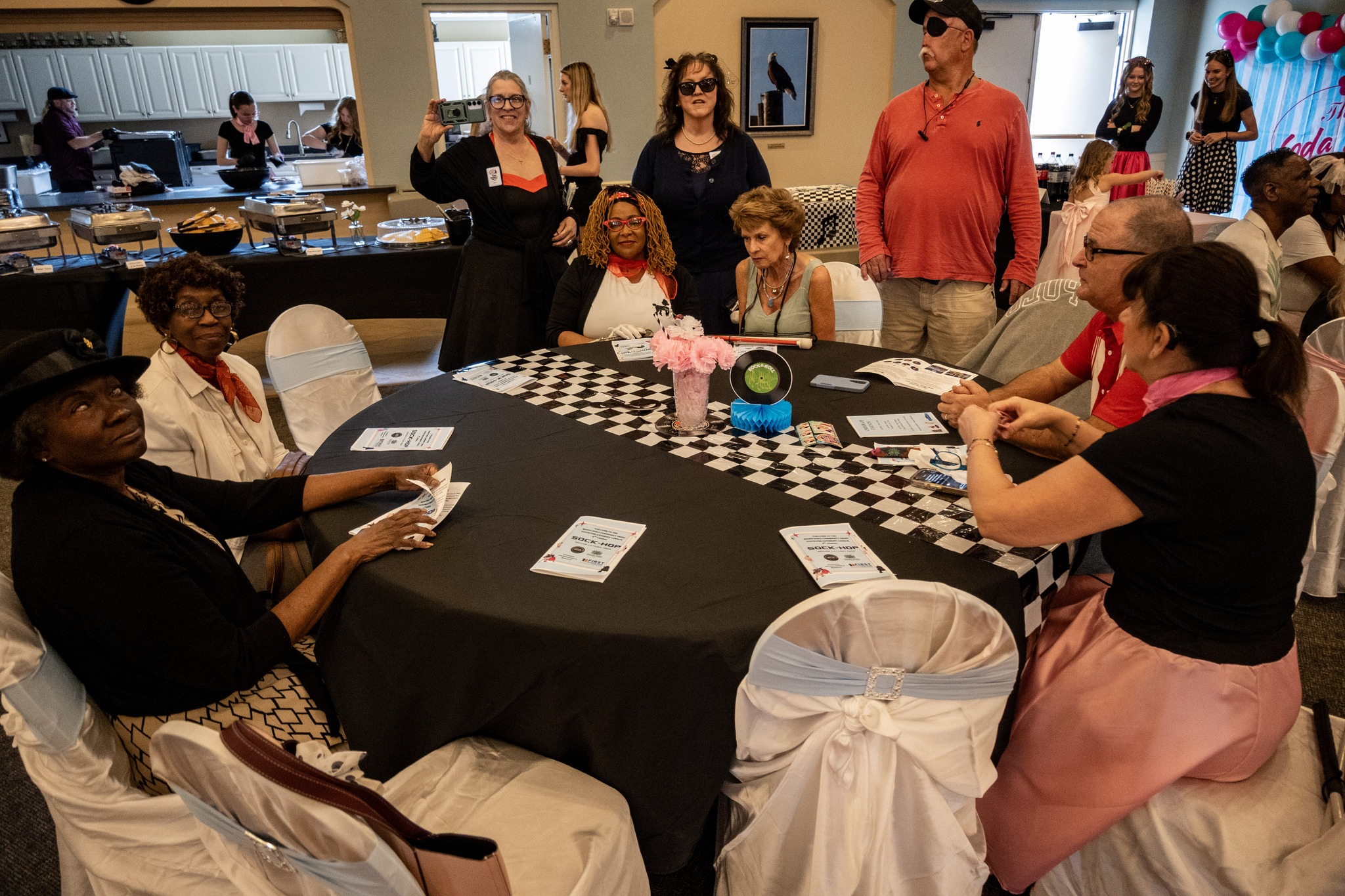 Attendees sit at one of the tables, smiling and talking during the Senior Citizens Prom. The table is decorated with a black and white checkered runner and pink flowers.
