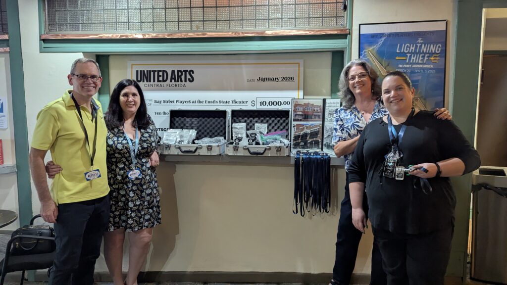 Staff from the Central Florida Audio Description Initiative stand by their set up in the theater. Two people on each side frame the table set up with two cases of the devices used for audio description. 