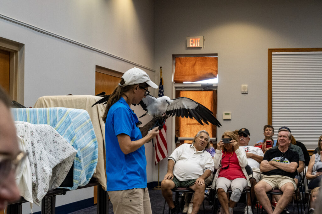 A volunteer from the Avian Reconditioning Center (ARC) holds a swallow-tailed kite on a gloved hand and speaks to the audience. New Vision clients listen and take pictures in the background.
