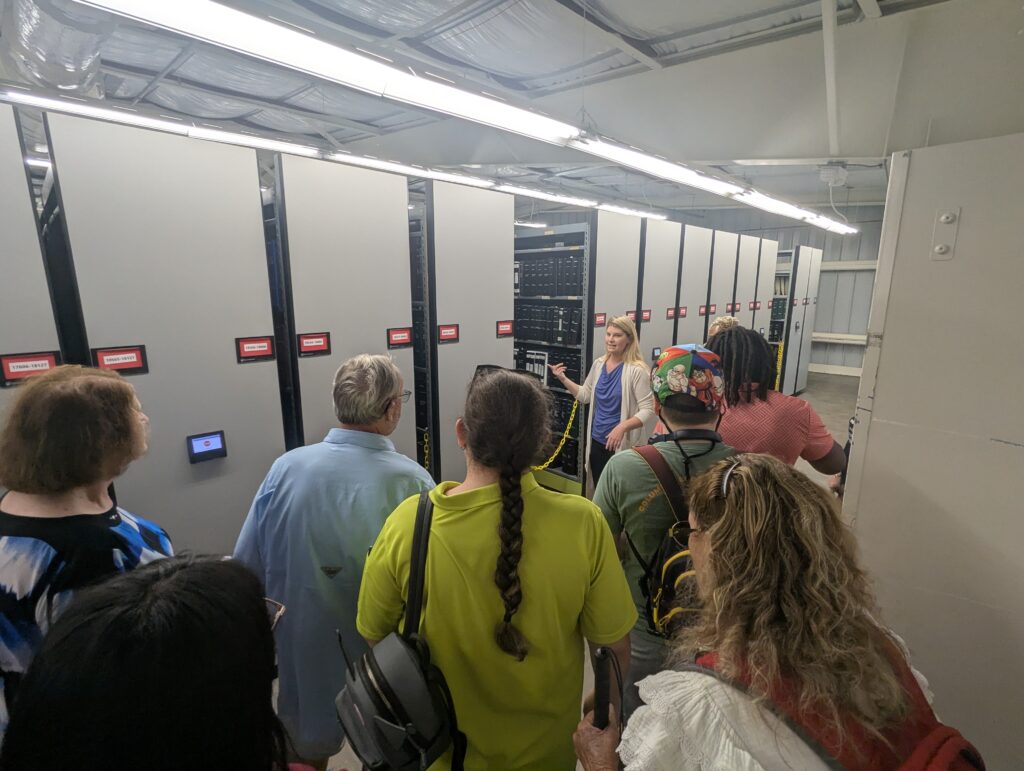 Teen and adult visitors from New Vision for Independence gather between rows of mobile library shelving while a staff member explains the Braille and Talking Book Library's collection.
