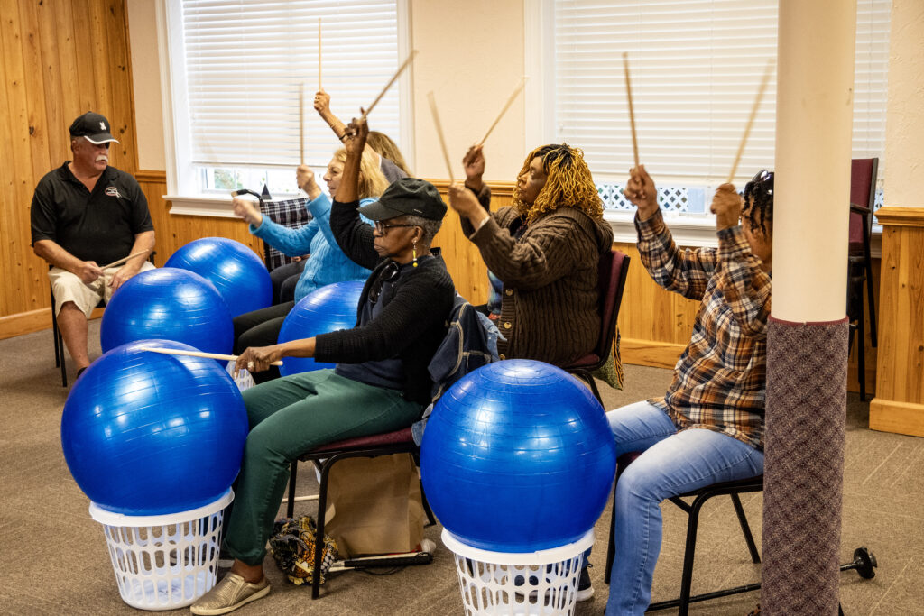A group of New Vision clients is seated, each with a bright blue exercise ball atop a small white laundry basket in front of them. They each have a pair of drumsticks, and are following along as Lisa instructs them on hitting the ball in various patterns. The group raises their drumsticks above their heads in tandem and clacks them together in time to the music. New Vision's Scott sits in the corner, waiting to assist.