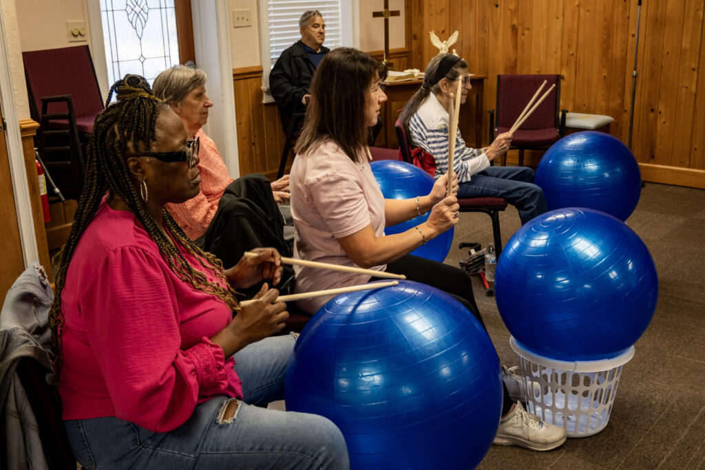 A group of New Vision clients is seated, each with a bright blue exercise ball atop a small white laundry basket in front of them. They are lifting up their drumsticks to hit the ball.