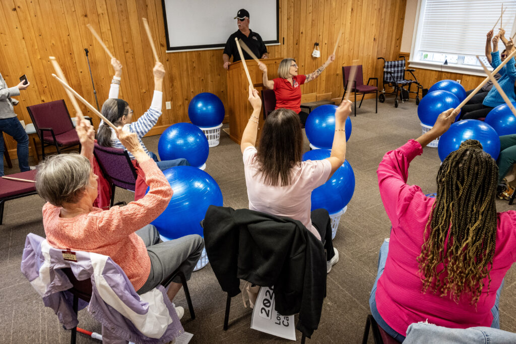 Lisa instructs the group to clack their drumsticks together in an overhead motion. Everyone is lifting their drumsticks to follow along.