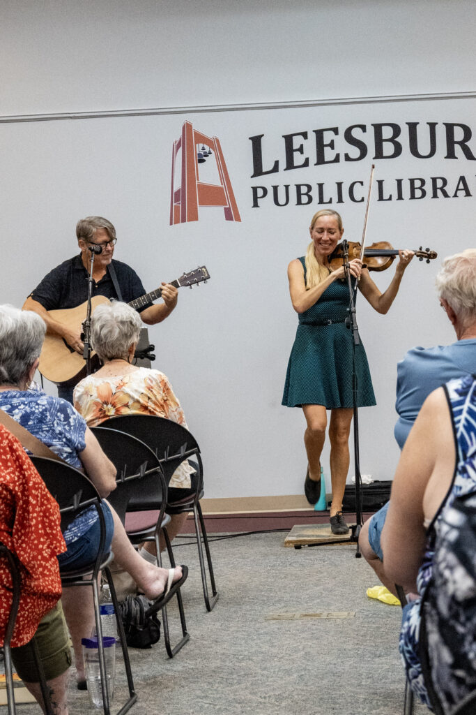 Looking down the aisle between rows of guests, two musicians, one on a guitar and one on a fiddle, play at the Leesburg Library's Celtic music performance. The library's logo is seen behind them on the wall
