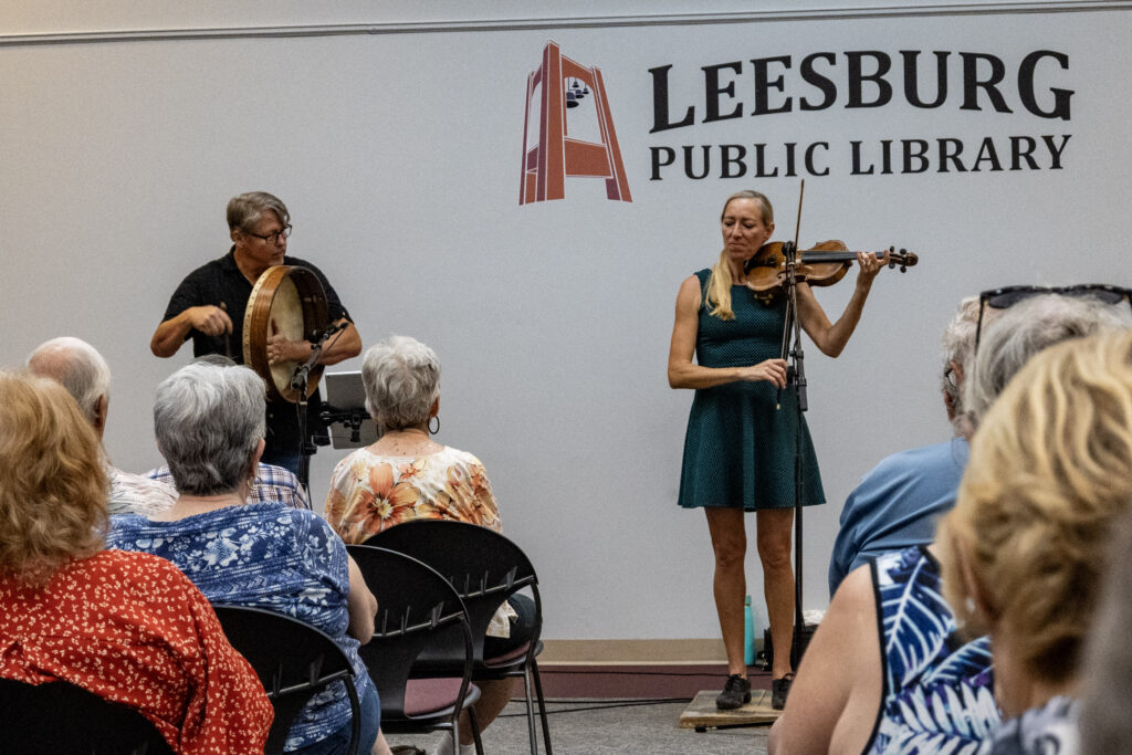 Looking down the aisle between rows of guests, two musicians, one on a bodhrán drum and one on a fiddle, play at the Leesburg Library's Celtic music performance. The library's logo is seen behind them on the wall.