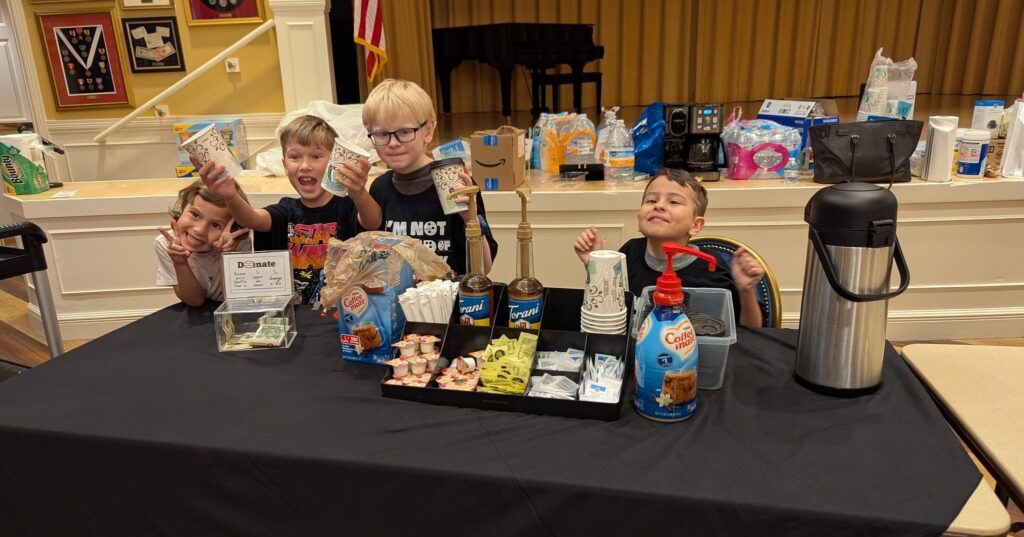 Four of the kids pose for a photo at the Low Vision Expo. On the left, one holds up two peace signs, the two kids in the middle hold up coffee cups, and on the end, one kid pumps his fists into the air.
