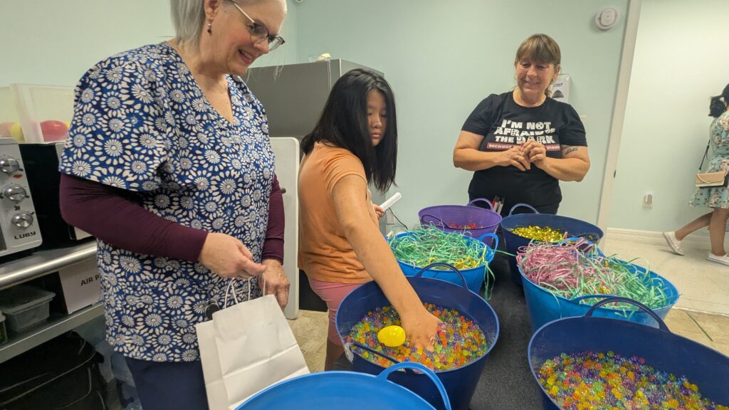 A girl reaches into a bucket filled with water beads, searching for Easter eggs. Other sensory buckets are lined up on the table. An aid stands next to her, and New Vision's Rebecca waits to refill the buckets with more eggs
