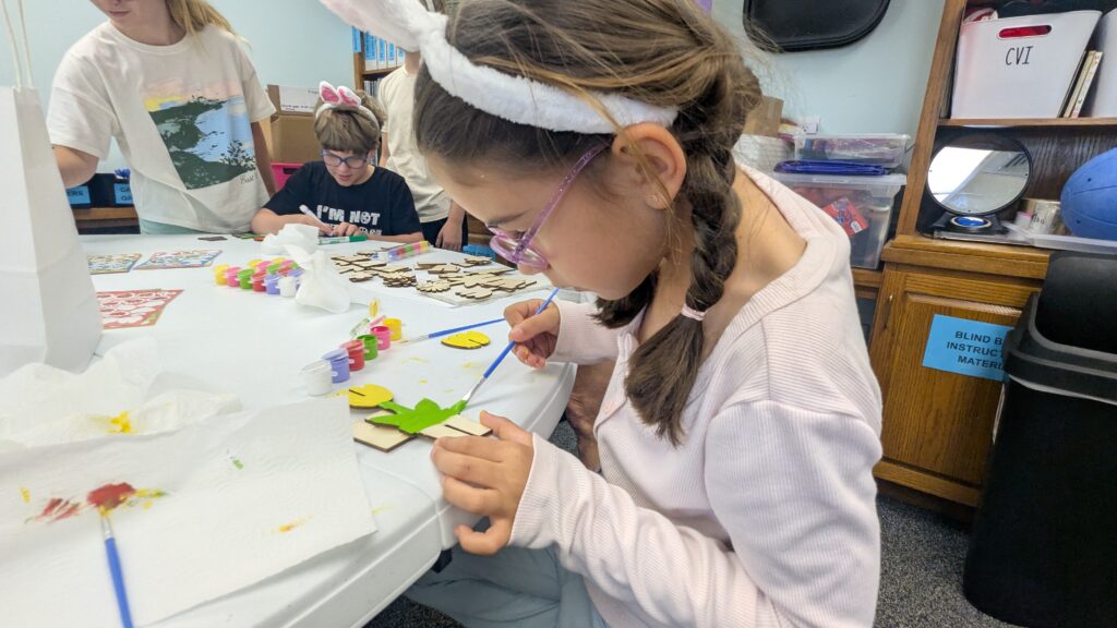A girl in bunny ears paints a wooden tulip with yellow paint.