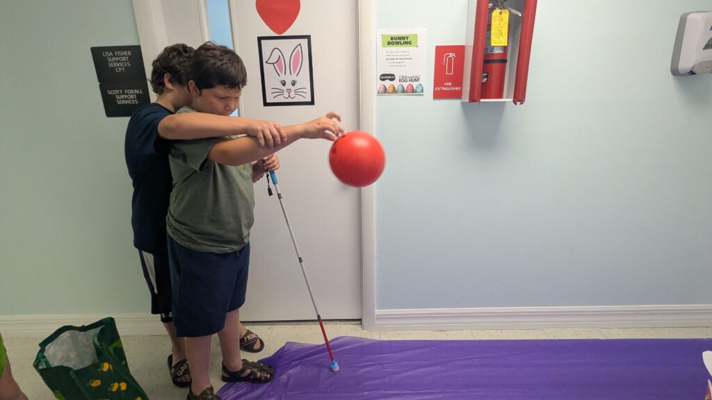 A boy using a white cane rolls a red plastic bowling ball down a lane with guidance from his brother.