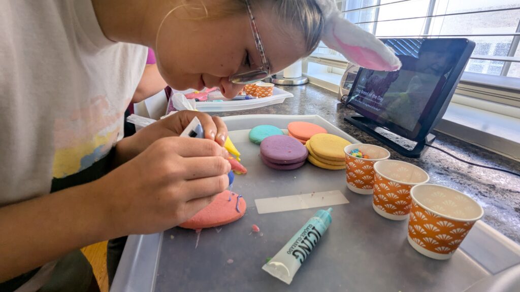 A girl in bunny ears ices a pink egg-shaped cookie with blue icing.