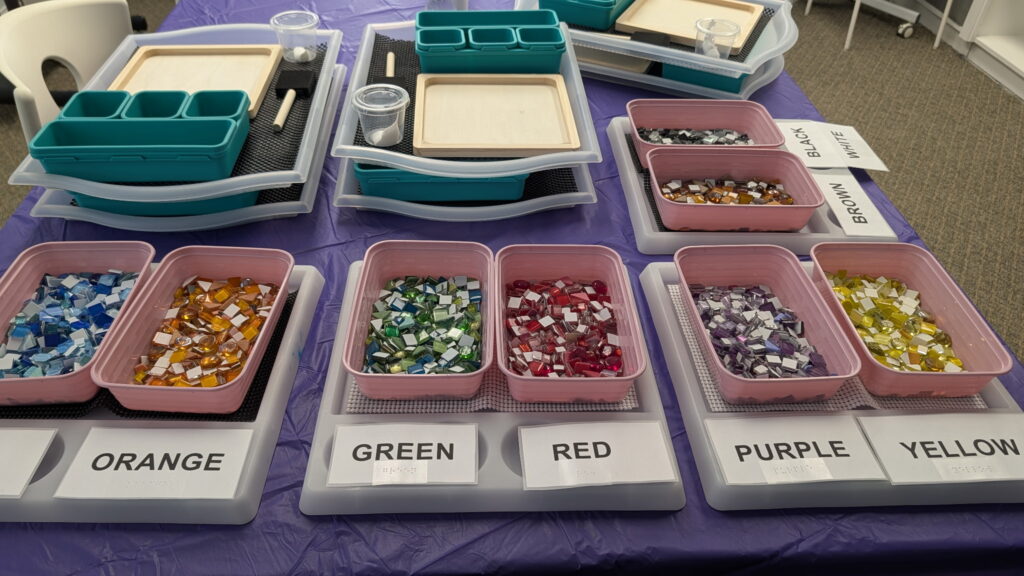 Bins of sorted tiles are labelled in large print and Braille and set up on a worktable. Stacked behind them are individual kits on trays for clients to use.
