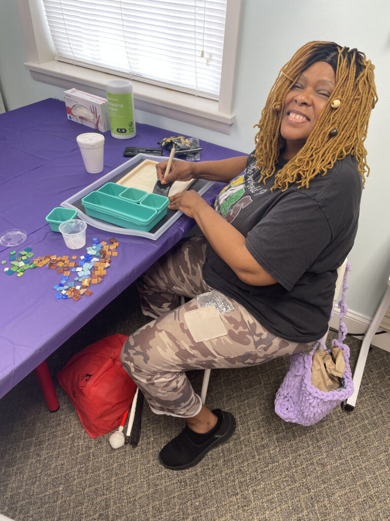 Wanda, board chair of New Vision, works on her mosaic project at the worktable, and smiles for a picture.
