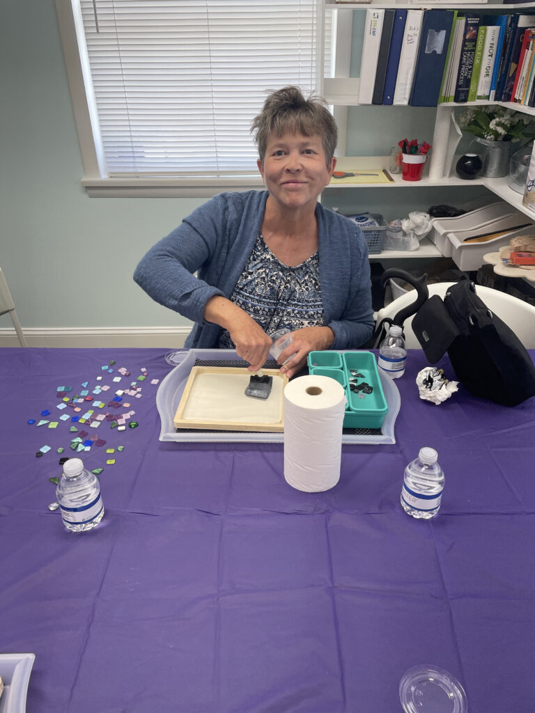 A New Vision client sits at a work table, spreading glue on the wooden frame with a sponge brush, and smiles for a picture.
