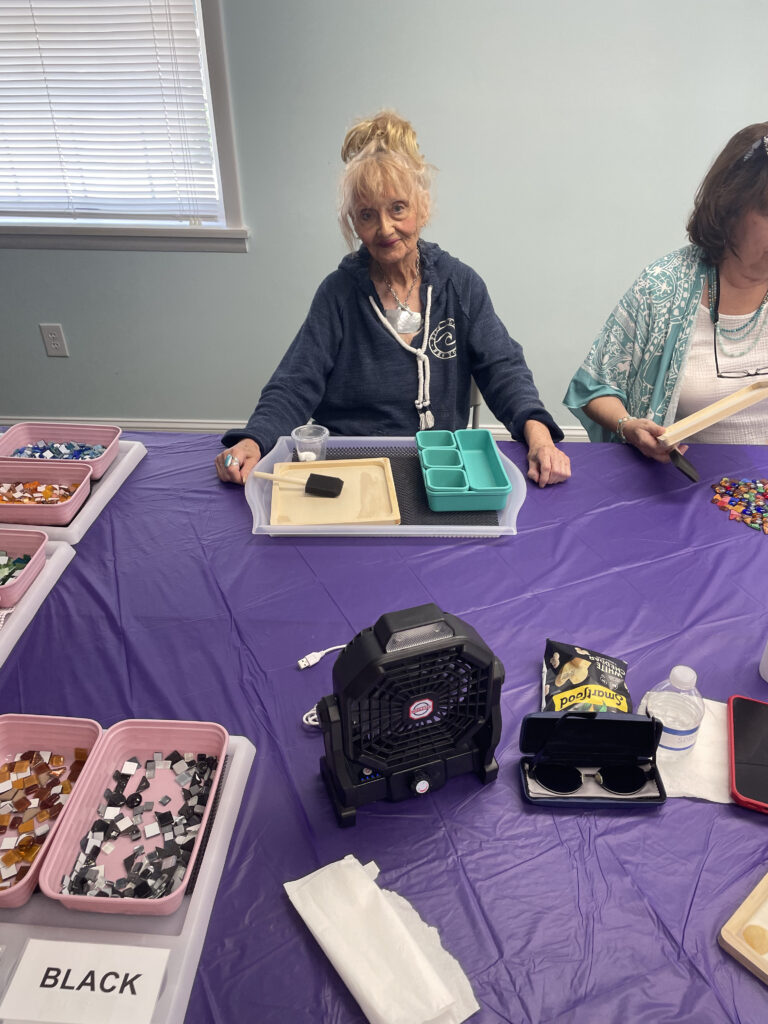 A New Vision client sits at a work table and smiles for the camera with her mosaic kit in front of her.

