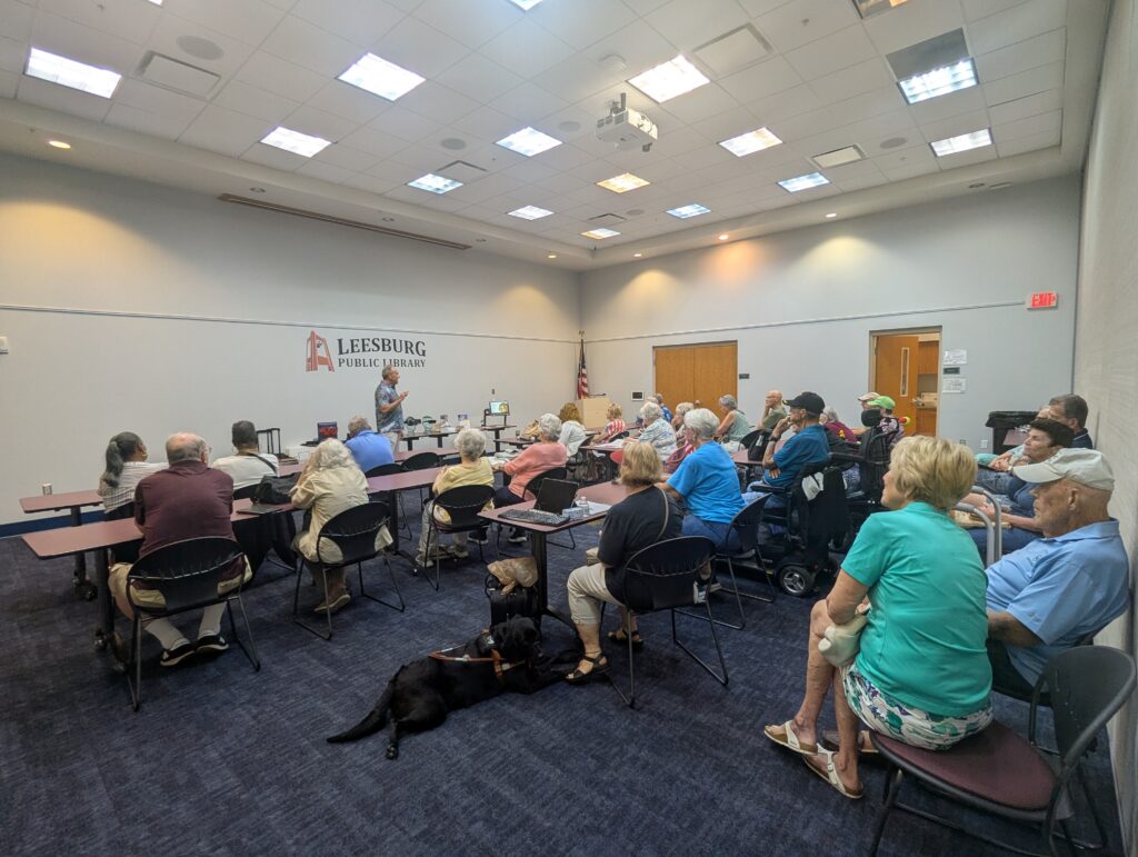 Bob, owner of The Magnification Co., stands at the front of a room in the Leesburg Library and address the crowd. Behind him are a row of magnification devices on a table.

