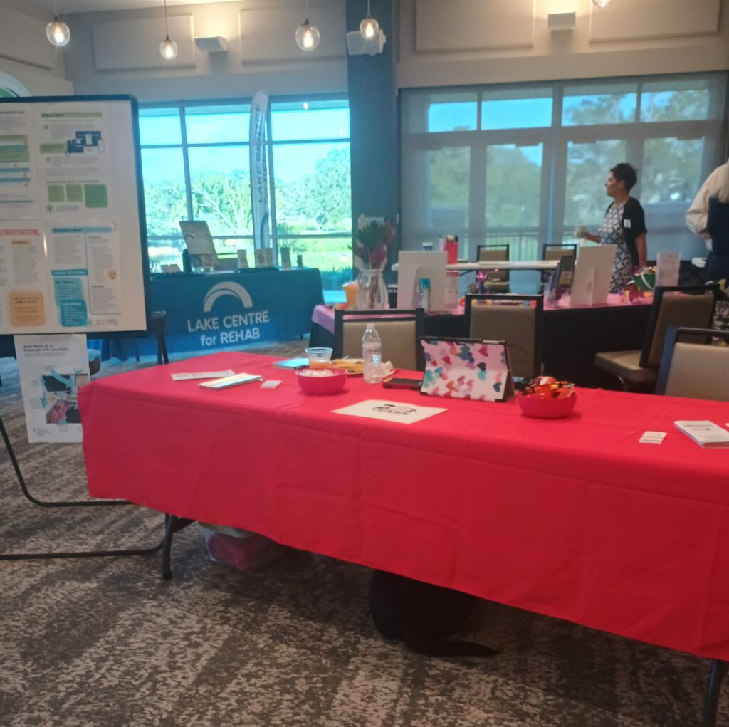 New Vision's table at the SHRM Summit is seen. Pamphlets are lain out on a bright red table cloth and there is a white board with printed flyers hung up.