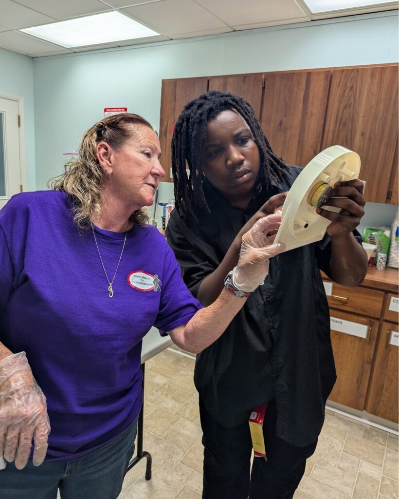 Jojo, Facilities & Safety Coordinator, stands beside Moses as they set a large print kitchen timer together. Jojo points out where to turn the dial to while making cake pops for New Vision's ribbon cutting.
