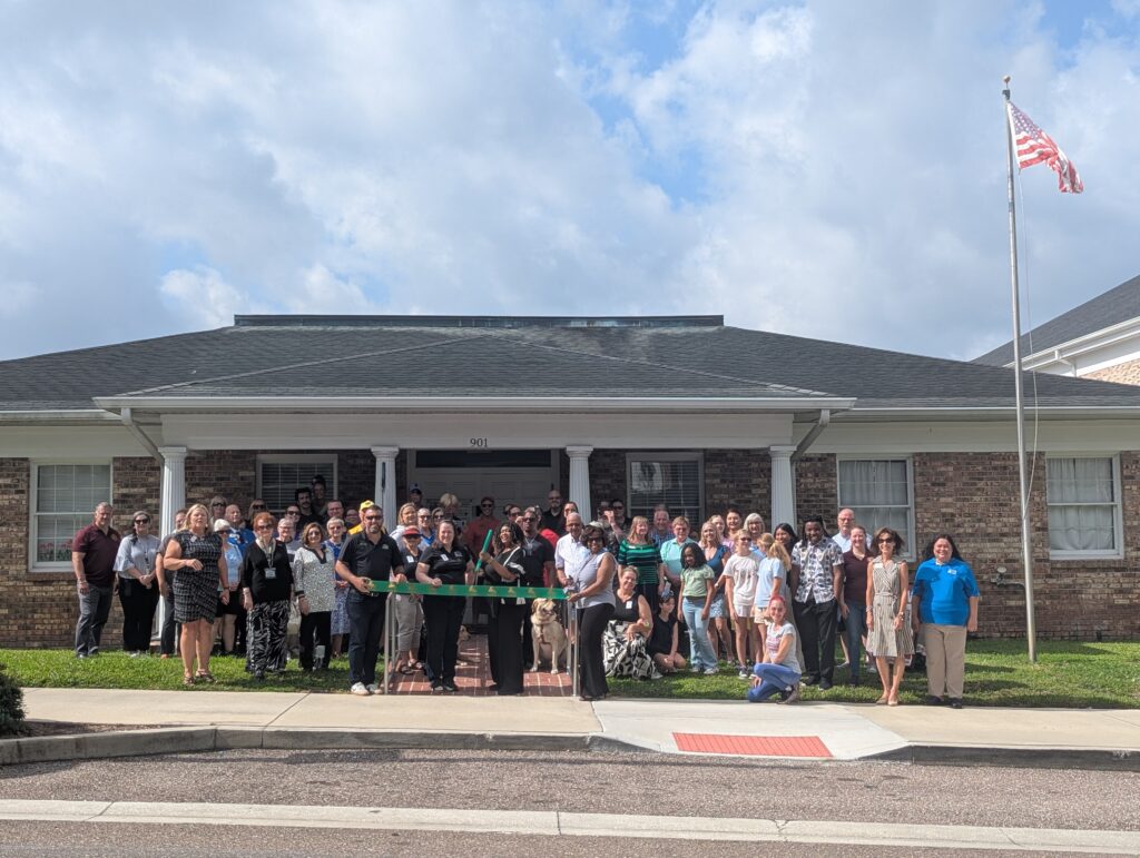 A group of more than 50 people stands in front of the New Vision for Independence building in downtown Leesburg for a ribbon-cutting ceremony. At the center, Board Chair Wanda McKenzie cuts a green ceremonial ribbon while Joyce Walters, Friendraiser & Donor Relations Coordinator, stands beside her. New Vision staff and board members are gathered on the front ramp, while community members, partners, supporters, and local leaders fill the lawn on either side. An American flag flies on a flagpole to the right of the building under a partly cloudy sky.