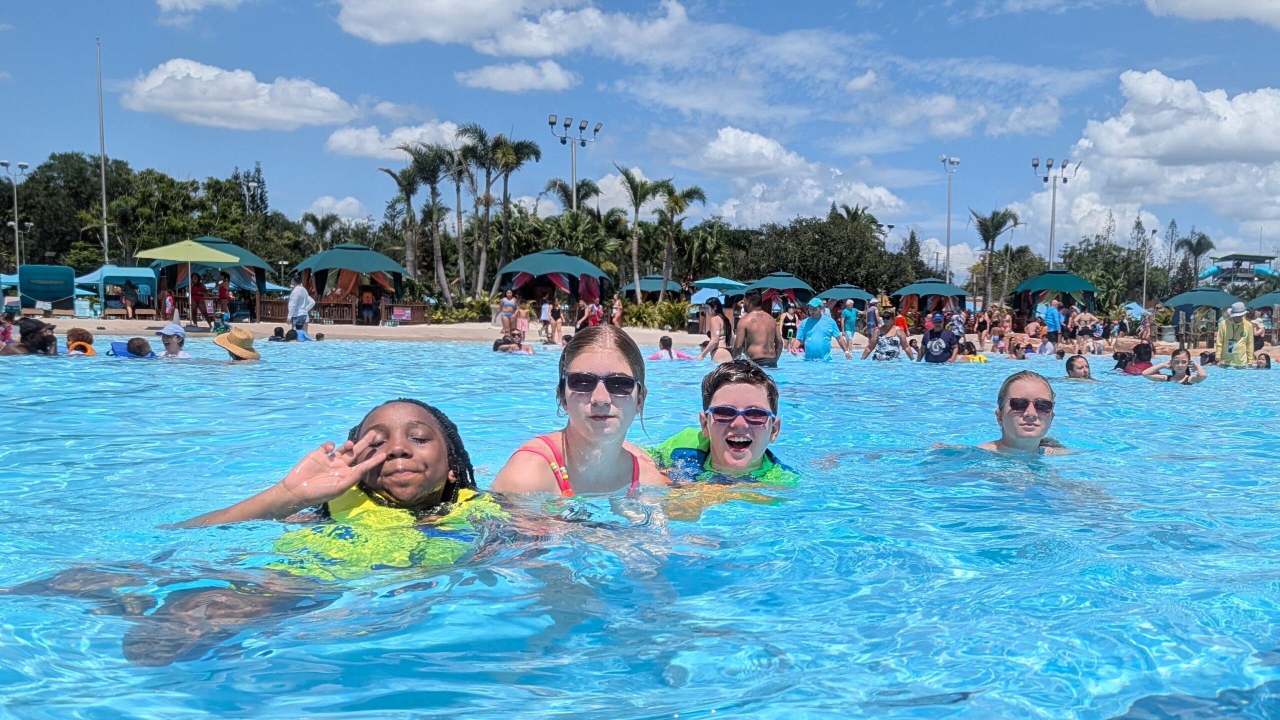 Four children from New Vision’s youth program pose together in a large swimming pool at Aquatica Orlando. One child waves toward the camera while another smiles broadly. Other swimmers, umbrellas, palm trees and water park features fill the sunny background.