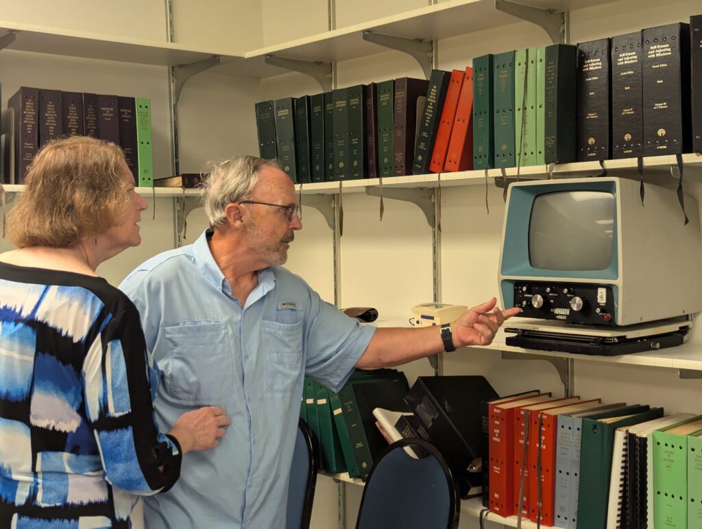Two visitors from New Vision for Independence examine a vintage video magnifier surrounded by shelves of Braille volumes during the tour of the Talking Books Library.