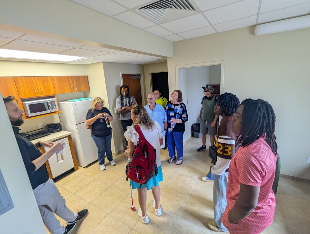 Teen and adult clients from New Vision for Independence gather in a furnished residential living area while touring the Daytona Beach training campus.