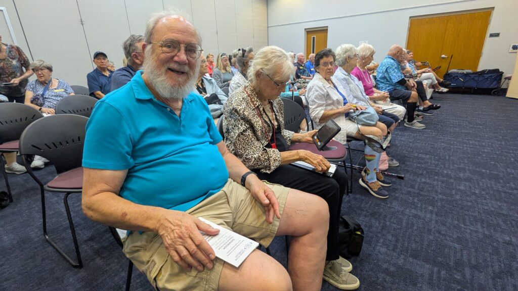 A smiling gentleman sits in the front row holding an event program while a woman next to him tests out a handheld magnifier at New Vision for Independence's Assistive Technology Expo at the Leesburg Public Library.