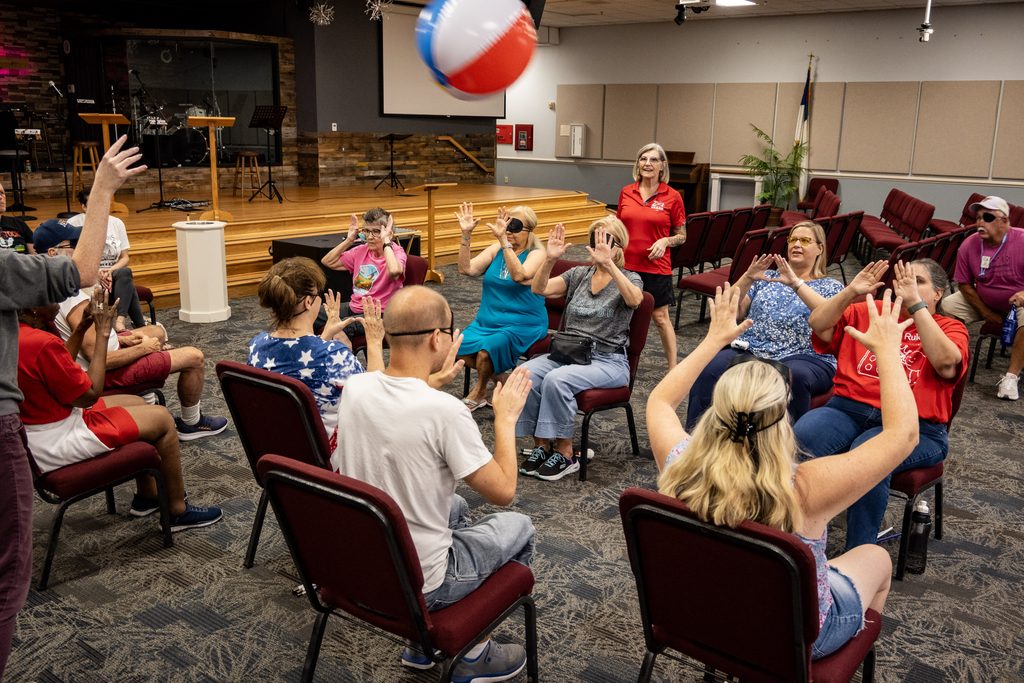 Participants sit in a circle with their hands raised as two colorful beach balls fly through the air during chair volleyball. New Vision staff stand among the group to help direct the game.