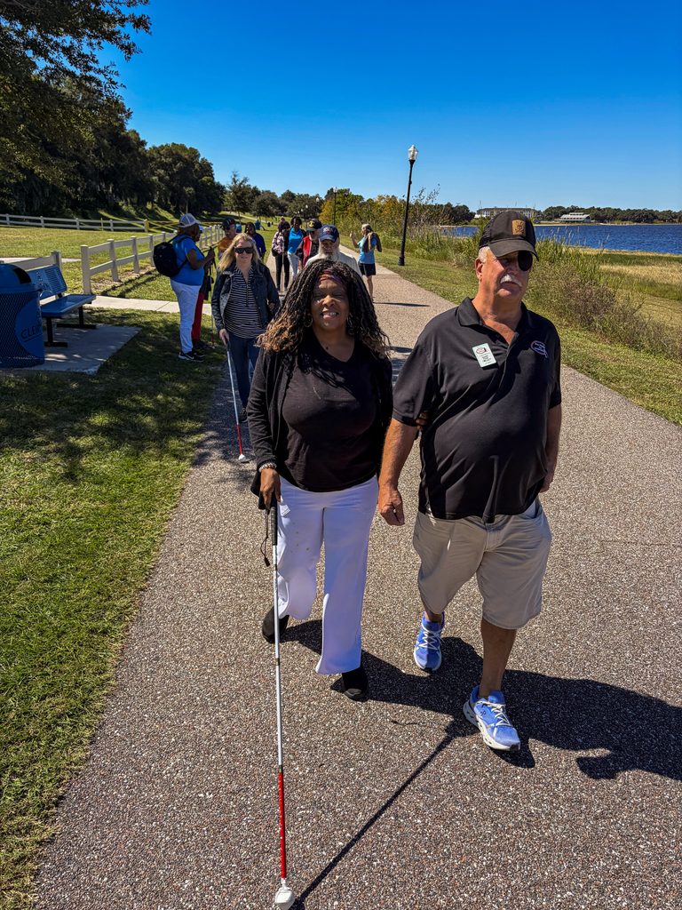 A woman wearing a black top and white pants walks along a waterfront path using a white cane while holding the arm of a man in a black polo shirt. Other participants follow behind them.