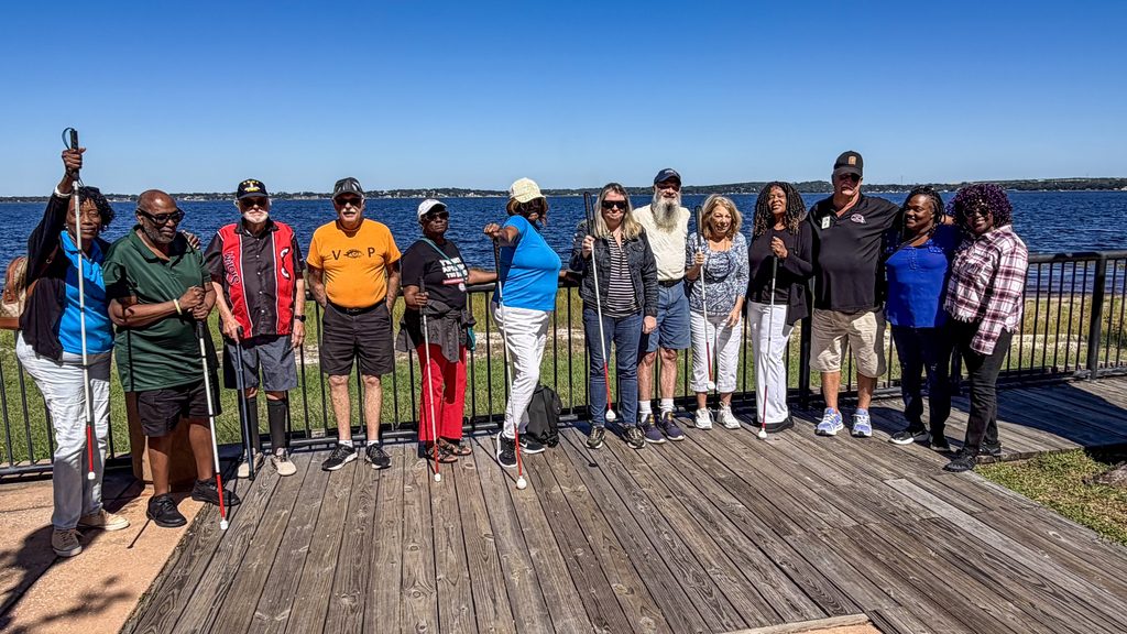 Thirteen participants pose together on a wooden waterfront boardwalk with a large lake behind them beneath a clear blue sky. The opposite shoreline in seen in the distance. Several participants hold white canes, and many are wearing hats and sunglasses.